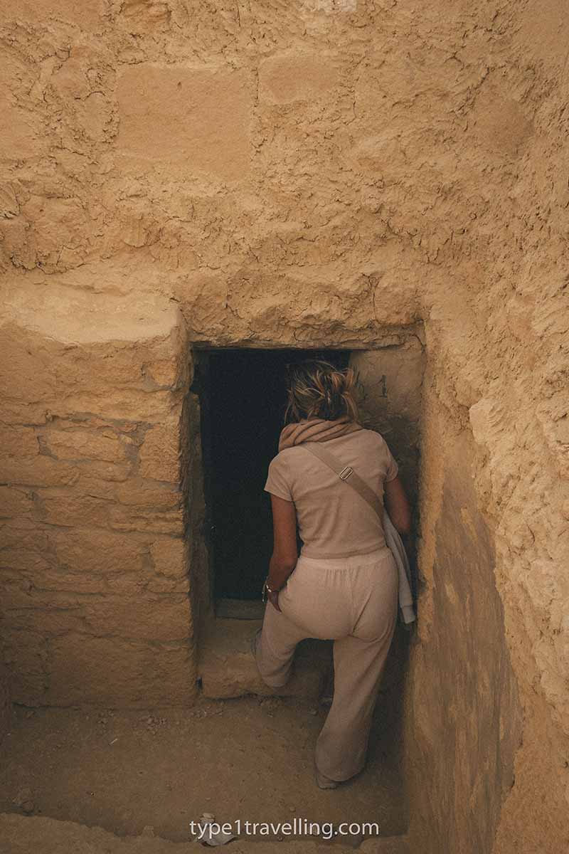 A woman stepping inside a small doorway leading into a dark tomb.