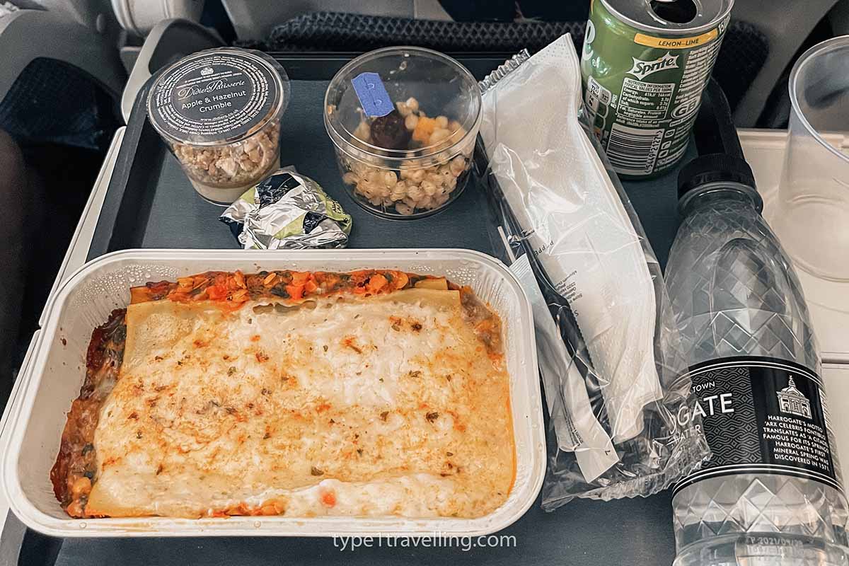 A tray with plastic cutlery and an airline meal of lasagne, apple and hazelnut crumble, quinoa salad, can of Sprite, and bottle of water.