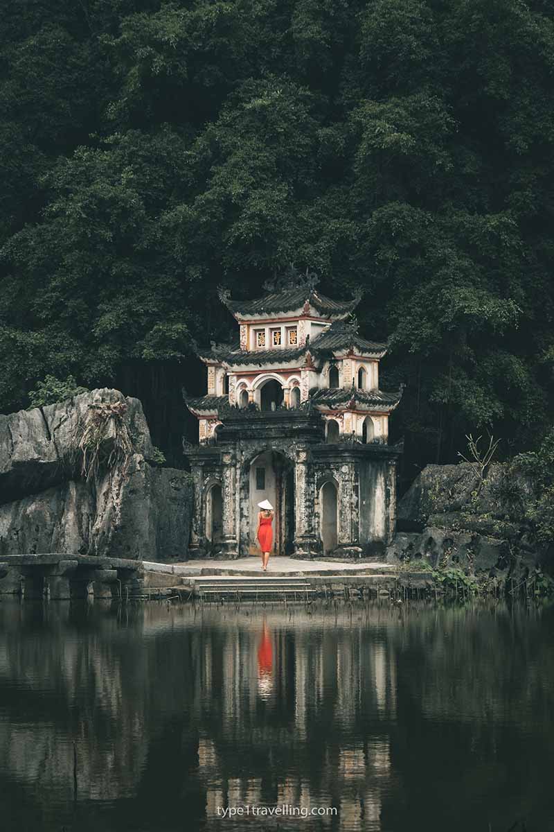 A woman wearing a red dress and non la hat walking towards Bich Dong Pagoda in Tam Coc, Vietnam.