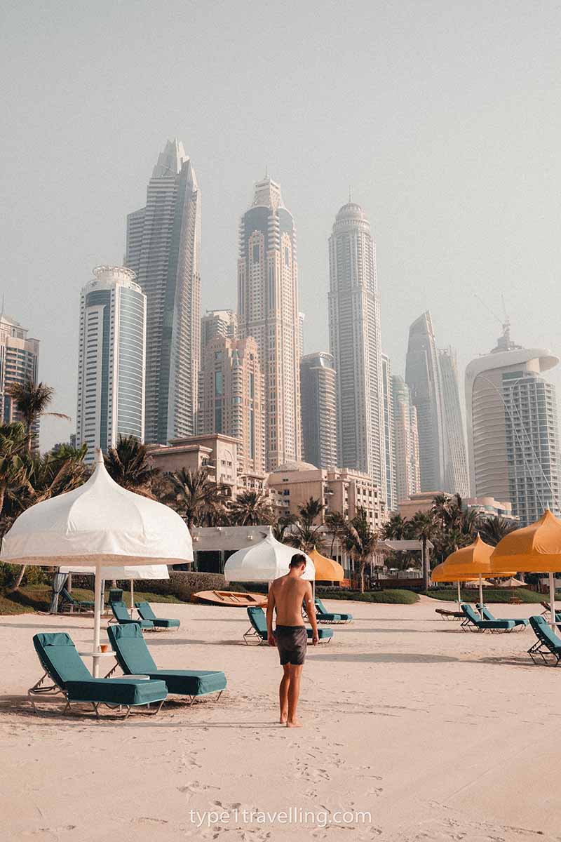 A man walking along a sandy beach in front of the skyline of Dubai Marina.