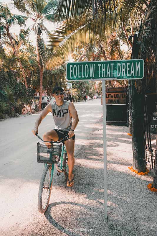 A man cycling past a sign that reads "follow that dream" along a palm tree-lined road.