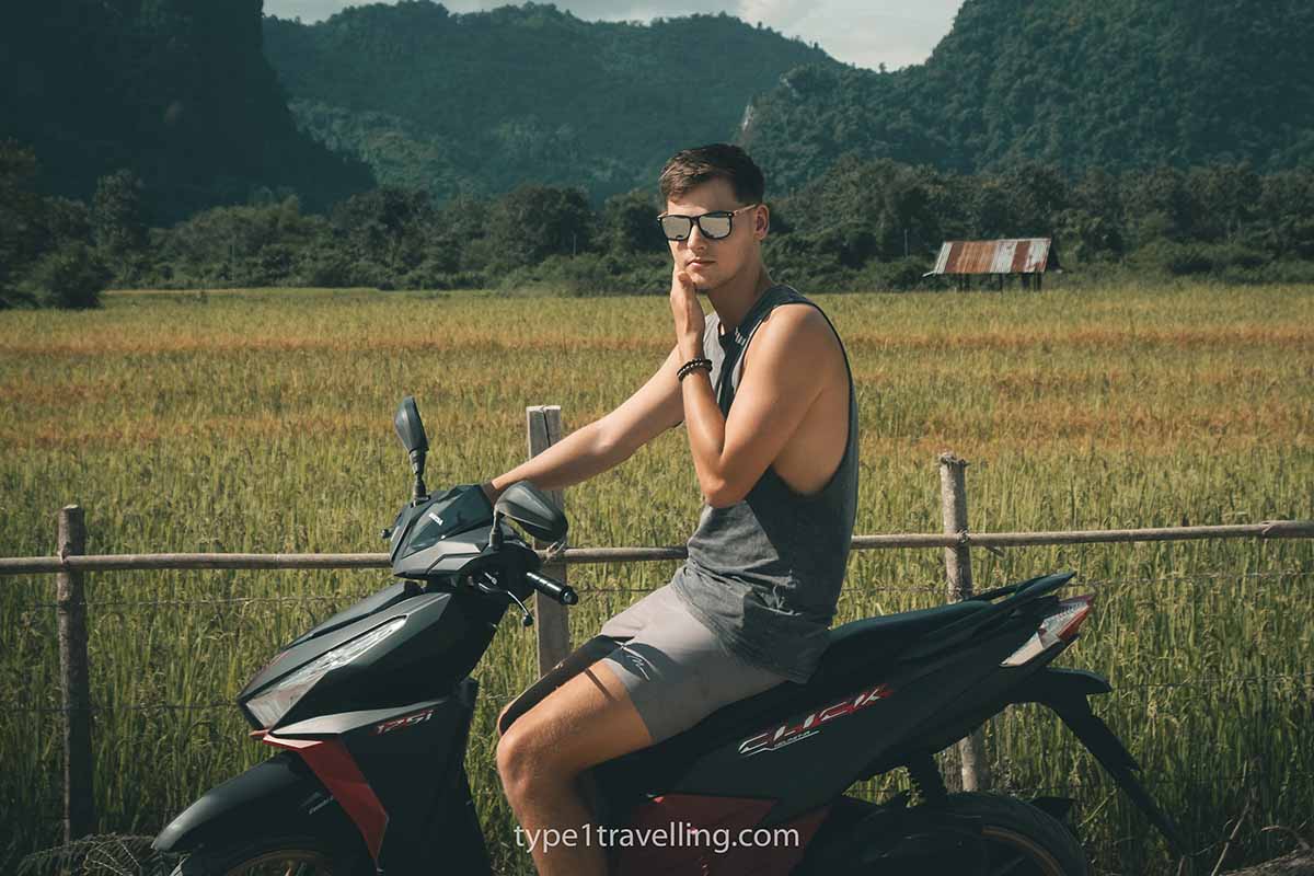 A man sitting on a parked scooter next to a rice field in the Laos countryside.