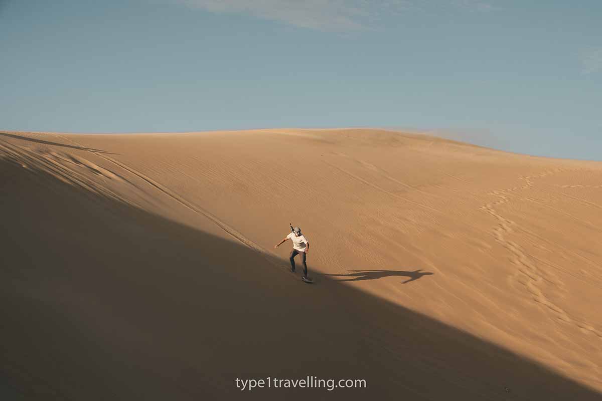 A person sand boarding down a large orange sand dune that is partly in shadow.