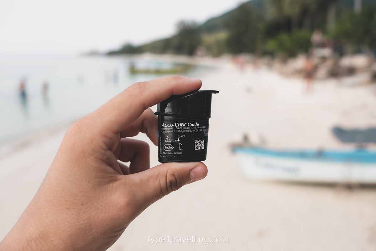 A person's hand holding a box of Accu-chek Guide test strips on a beach.