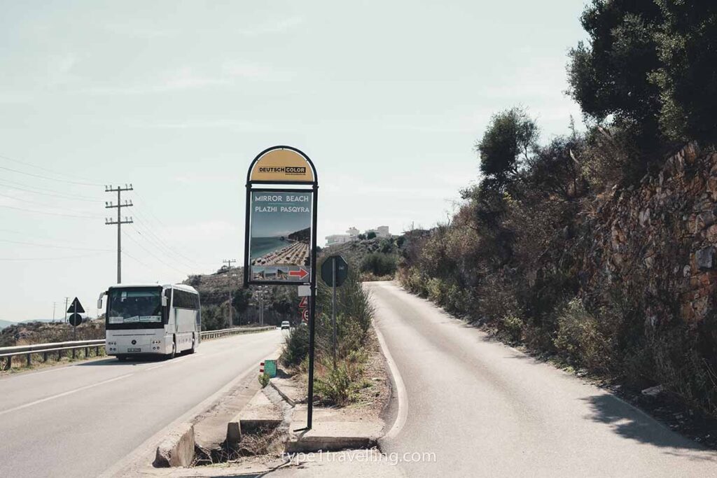 mirror-beach-turn-off A sign which reads "Mirror Beach Plazhi Pasqyra" and points to a single track road that runs parallel to the single carriageway.