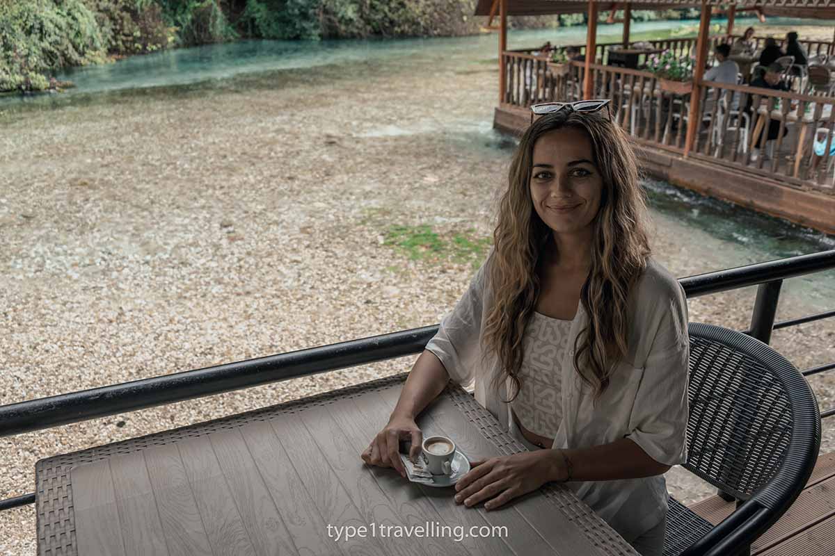 A woman smiling while sitting with a coffee at a table next to the Bistrica River.