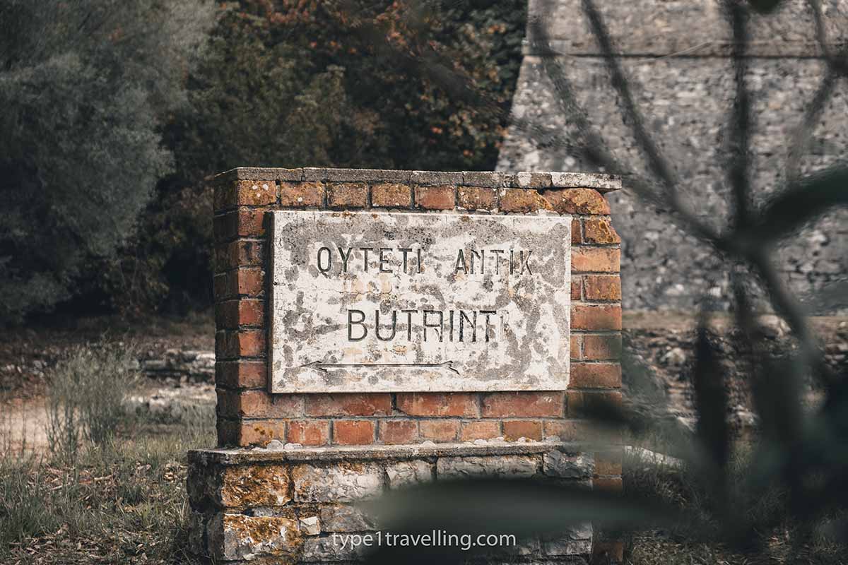 butrint-sign A worn stone sign attached to a red brick wall that reads "OYTETI ANTIK BUTRINT".