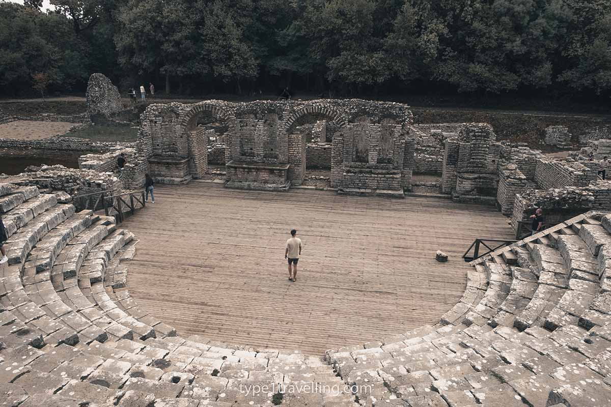 butrint-theatre A man walking on the wooden platform of the ancient Butrint Theatre which is surrounded by stone ruins.