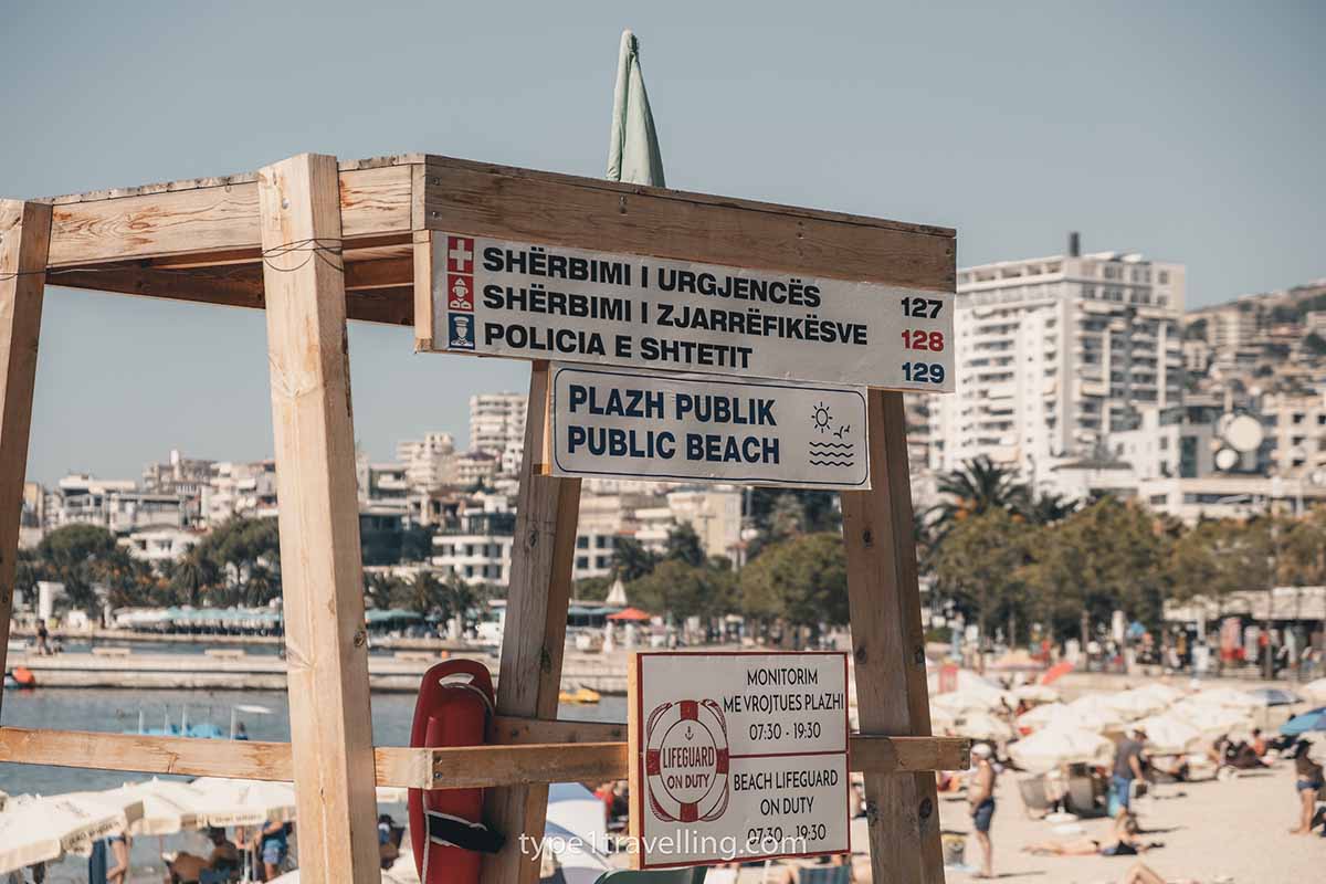 A wooden lifeguard tower which displays a sign which reads public beach in English and Albanian.