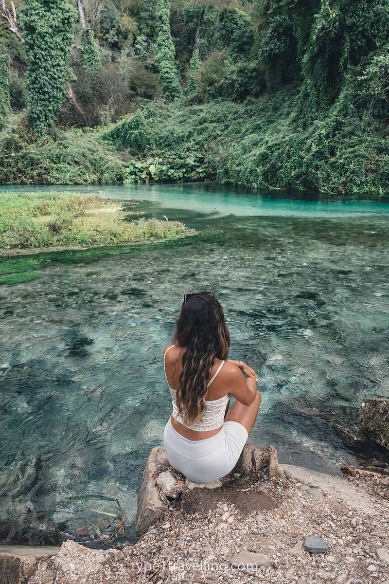 A woman sitting on a rock next to a shallow, clear-water river which is surrounded by lush greenery.