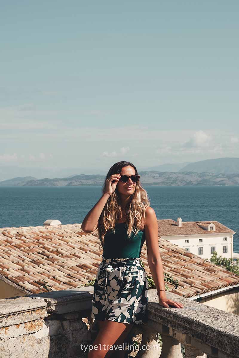 Awoman standing against a stone ledge with a red-tiled roof, sea, and mountains in the background.