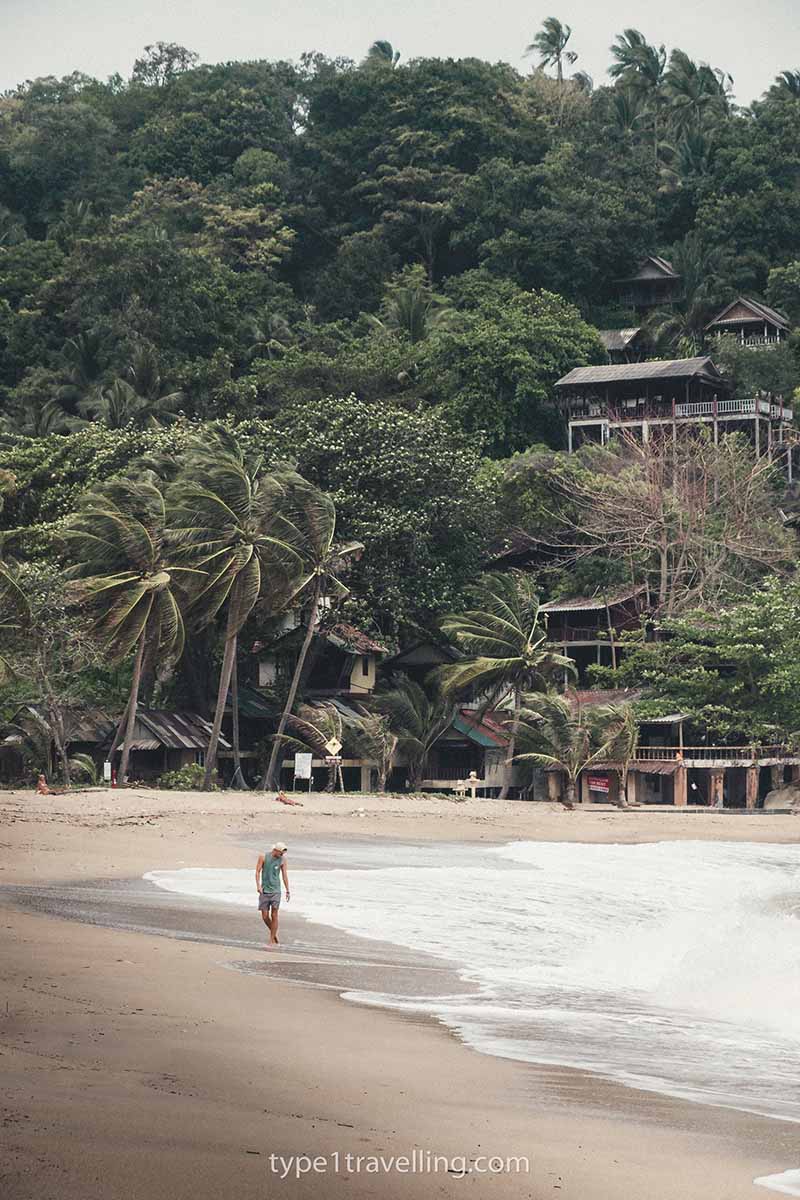 A person walking along the white water shoreline of Haad Sadet Beach with palm tree and raised bungalows in the background.