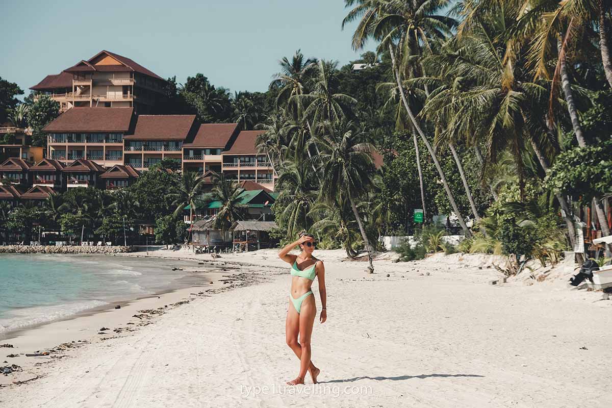 haad-yao-beach A woman walking on the white sand of Haad Yao Beach which is lined with overhanging palm trees with a beach resort in the background.
