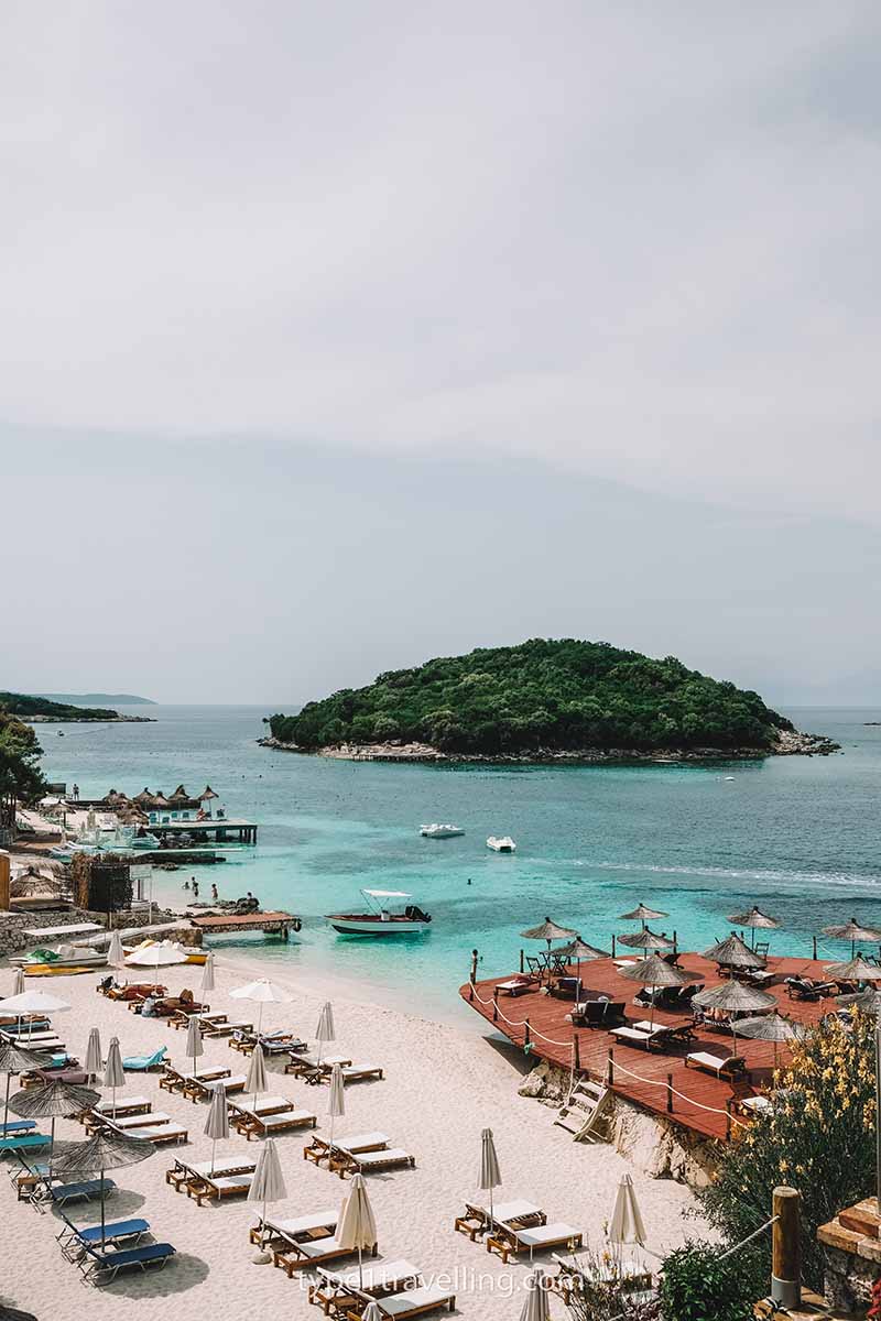 An empty beach lined with sun beds and closed beach umbrellas with a small island in the background.