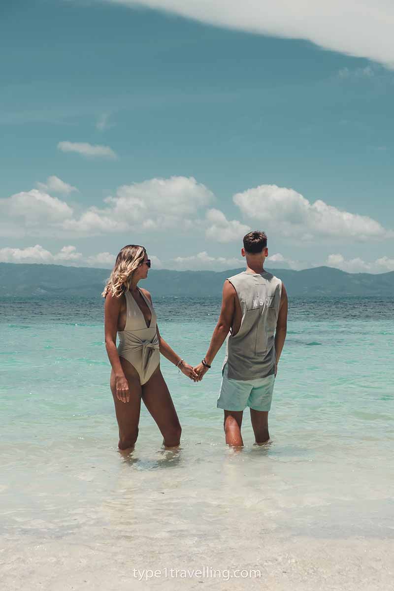 A couple holding hands and looking out to sea while standing knee-deep in clear turquoise water.