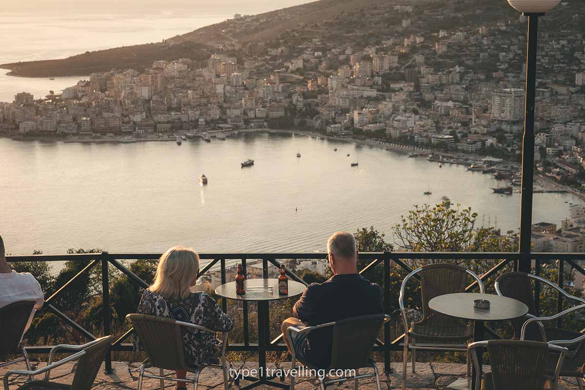 A couple sitting at an outdoor table with two bottles of beer looking over the bay of Saranda during sunset.