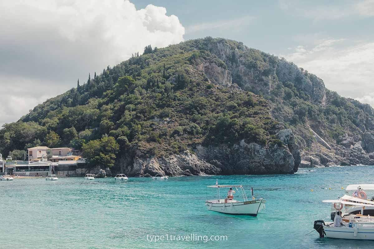 A person in a small boat on the turquoise water in front of a rocky, tree-covered headland.