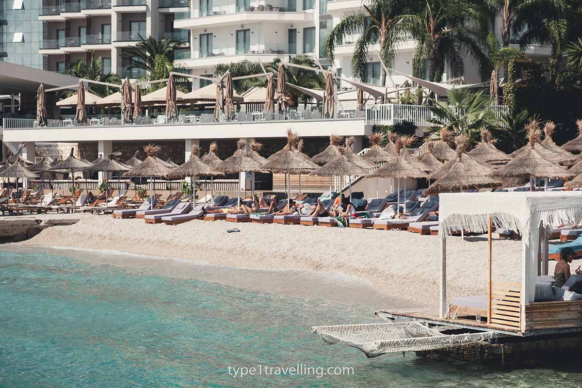The pebble shoreline of Plazhi Era backed by rows of sun loungers and parasols, and apartments.
