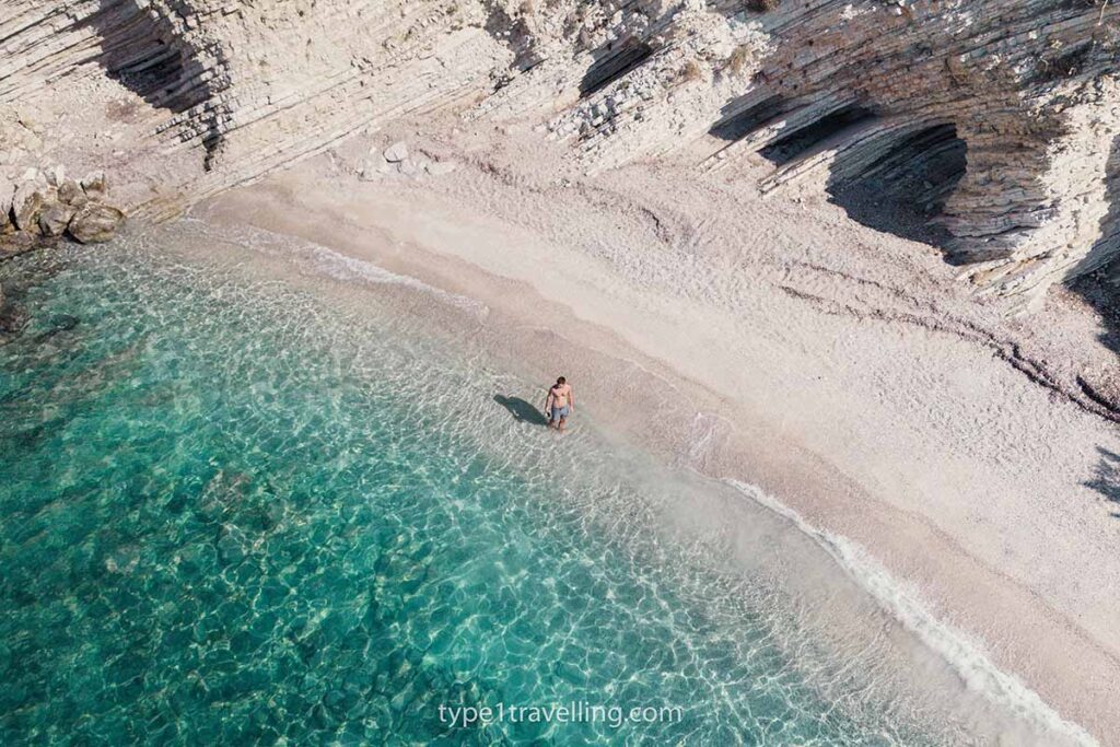 plazhi-i-pasqyrave-drone A person standing in the water close to the shore of Plazhi I Pasqyrave which is surrounded by eroded cliff-face.