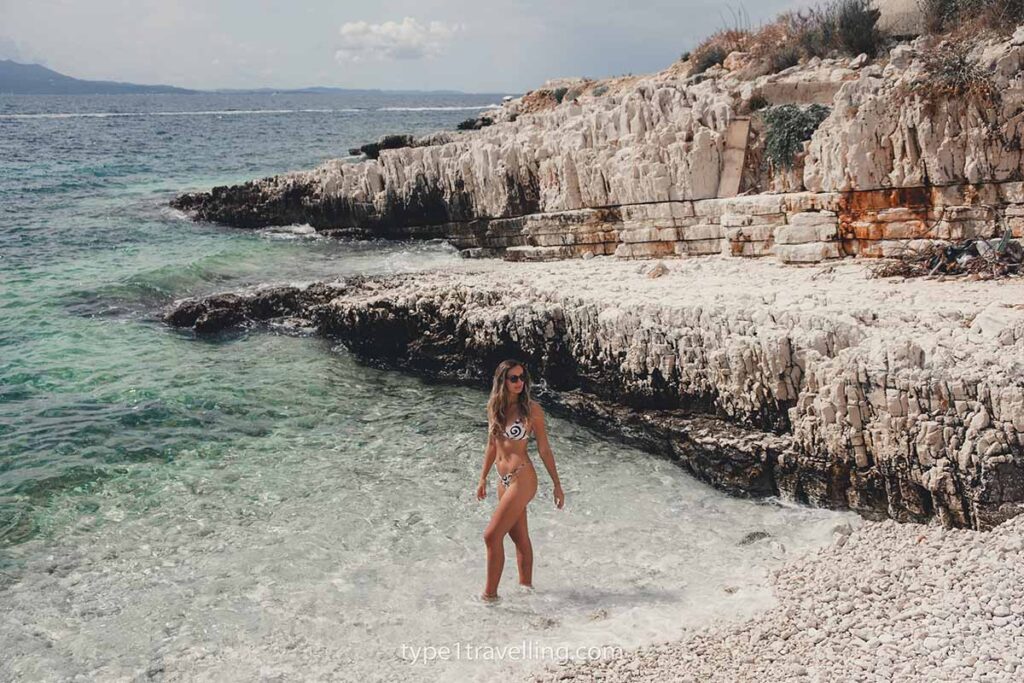 A woman paddling ankle deep in a small pebble cove which is enclosed by jagged rocks.