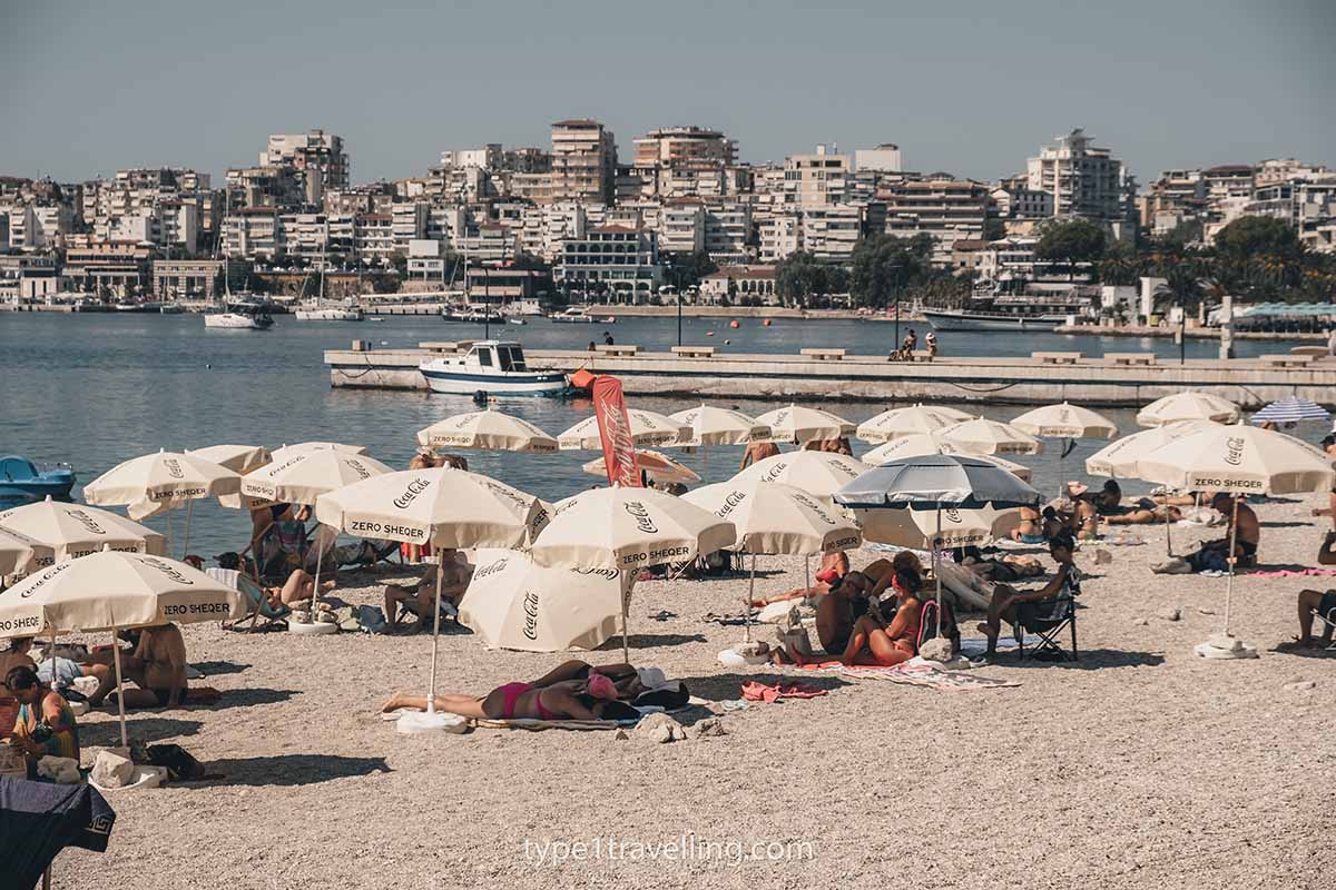People lying on beach towels under beige parasols on Saranda Public Beach.
