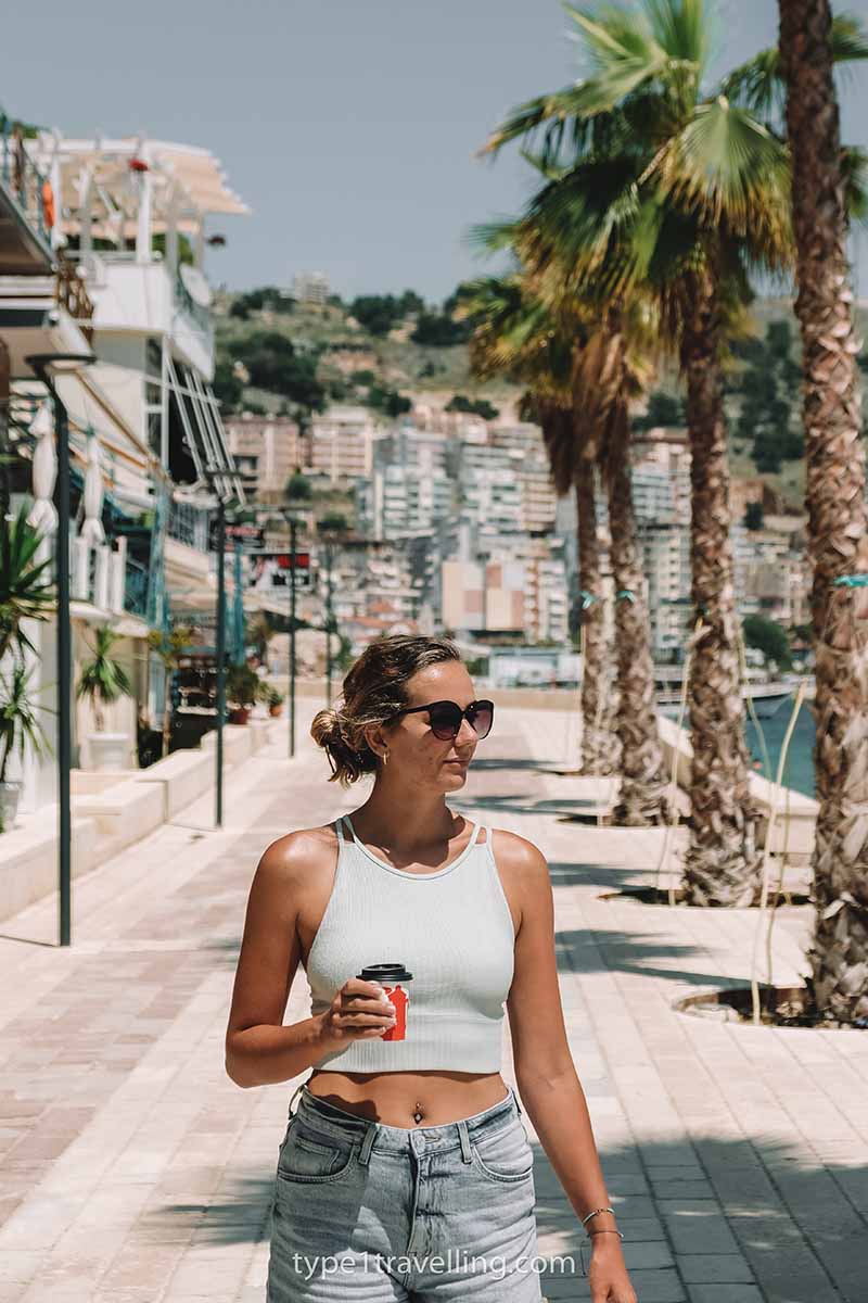 A woman with a take away coffee walking along Saranda Promenade which is lined with palm trees.