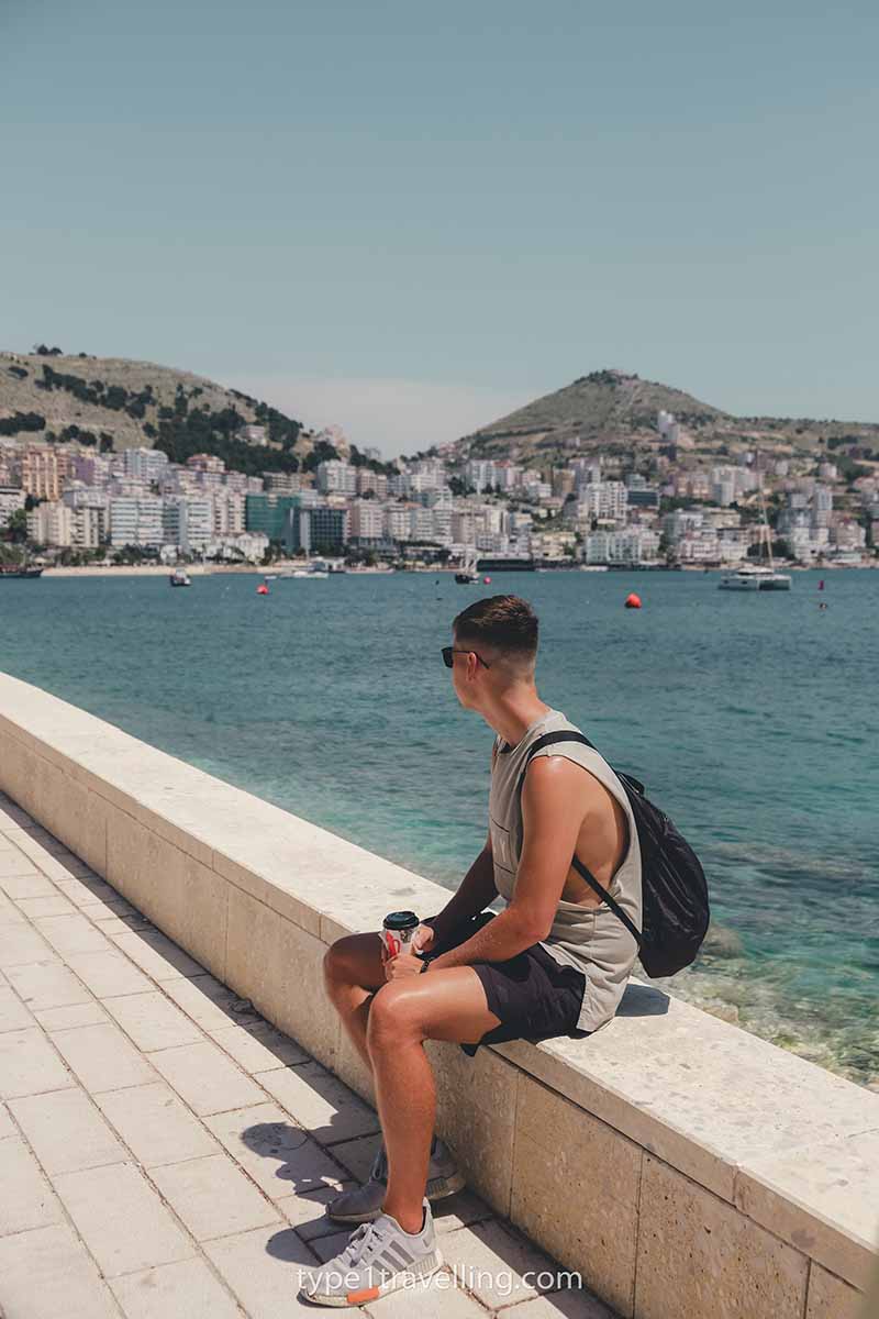 A man holding a coffee cup while sitting on the ledge of Saranda Promenade with the bay and apartment buildings in the background.