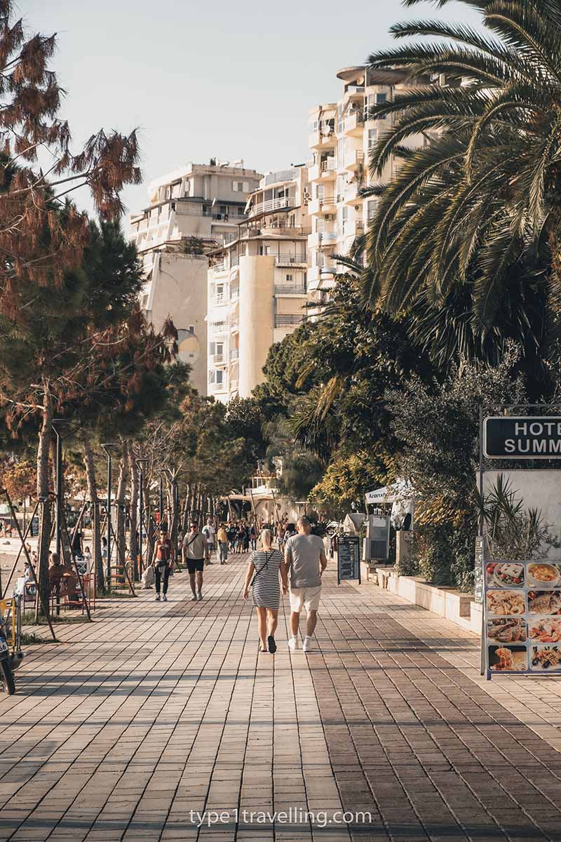A couple walking hand in hand along the Saranda Promenade which is lined with trees and apartment buildings.