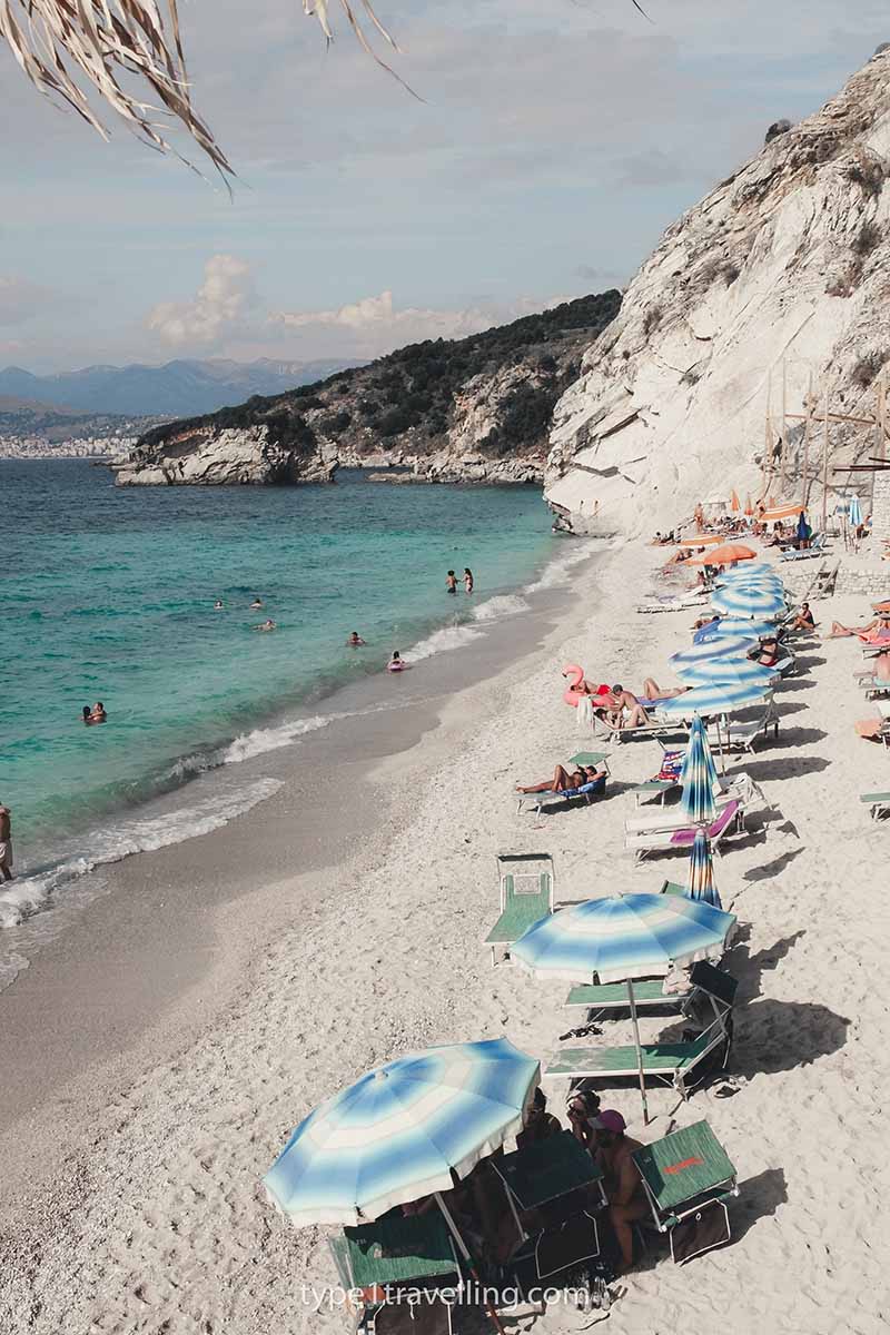 A row of blue and white parasols and sun loungers on the narrow strip of Shpella e Pellumbave Beach.