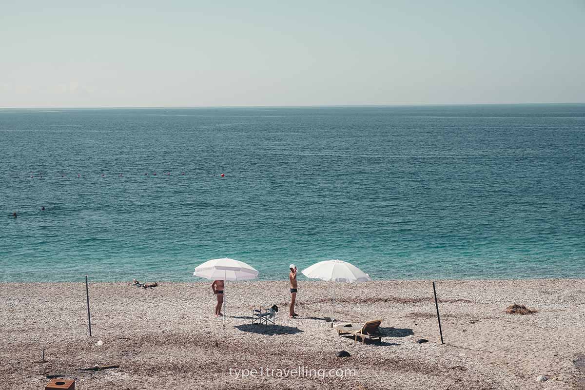 beach-in-himare Two people standing next to two parasols on a pebbles beach.