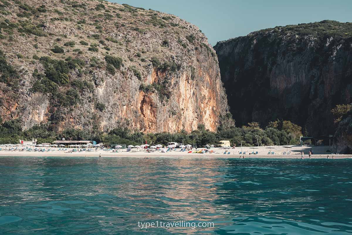 gjipe-beach-himare Lines of sun beds on Gjipe Beach with steep cliff faces and Gjipe Canyon in the background.
