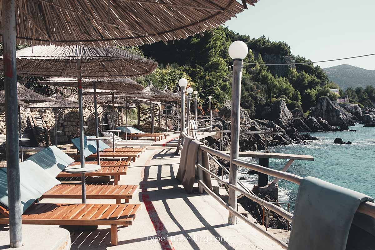 A line of wooden sun beds and straw umbrellas, and a wooden safety railing on the platform of Kico Beach.