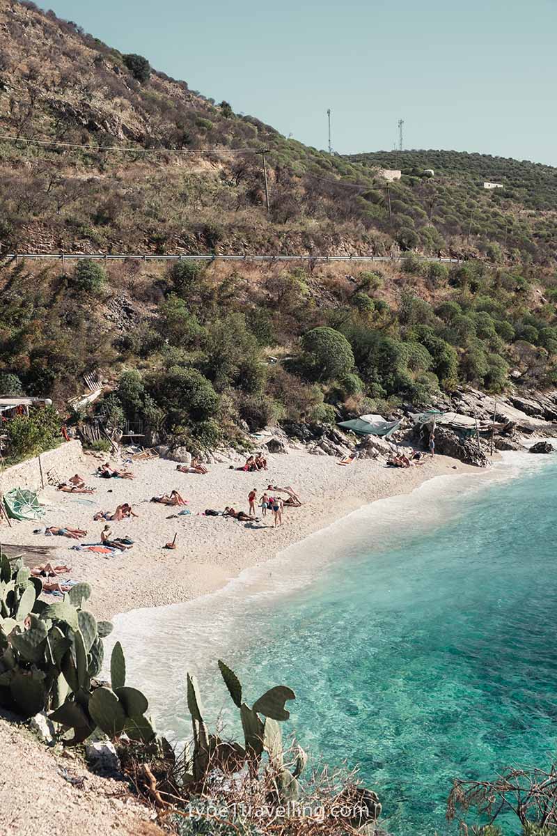 Several people sunbathing on Porto Palermo beach with large green hills in the background.