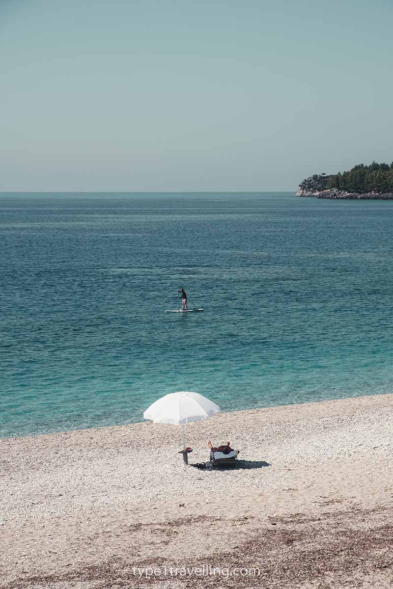 A single umbrella and sun bed on Potami Beach with a person paddle boarding in the background.
