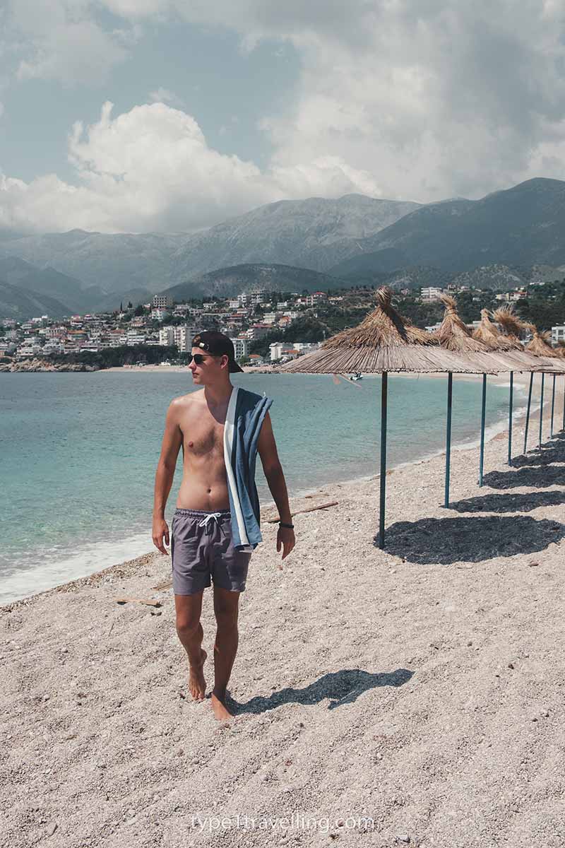 A man walking with a towel over his shoulder next to a line of straw beach umbrellas with the town of Himare and mountains in the background.