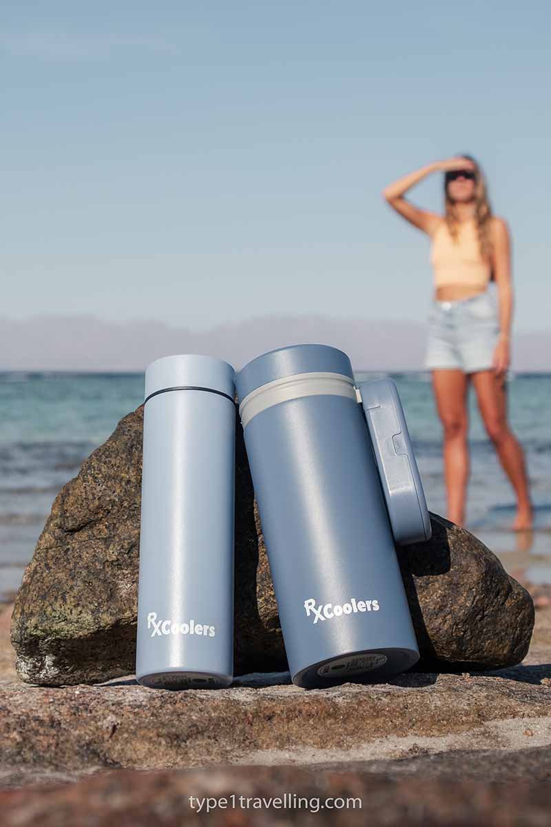 An RXCoolers Max and Mini model sitting against a rock while a woman walks along the beach shoreline in the background.