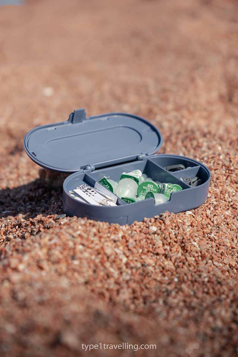 An open RXCoolers Max extra storage case sitting on sand with insulin needles and blood glucose test strips inside.