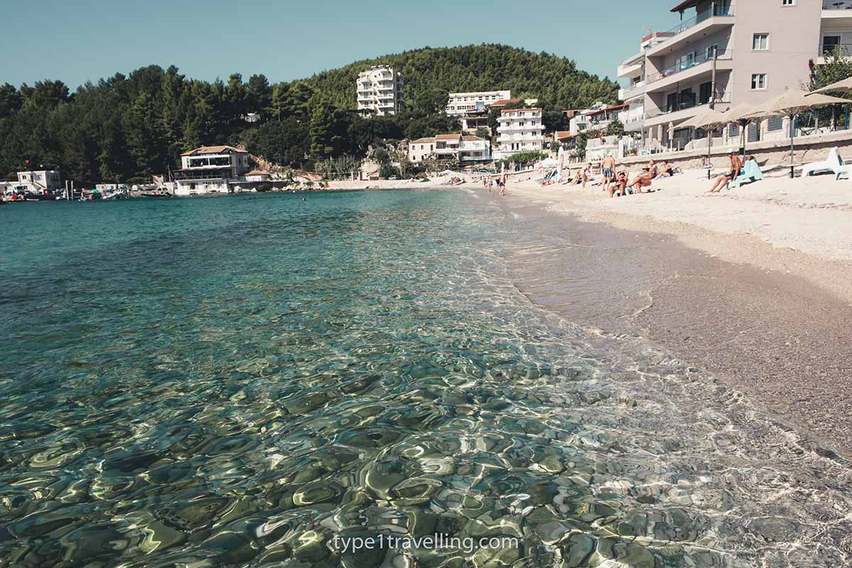The clear water and pebbles of Spile Beach with hotels and headland in the background.