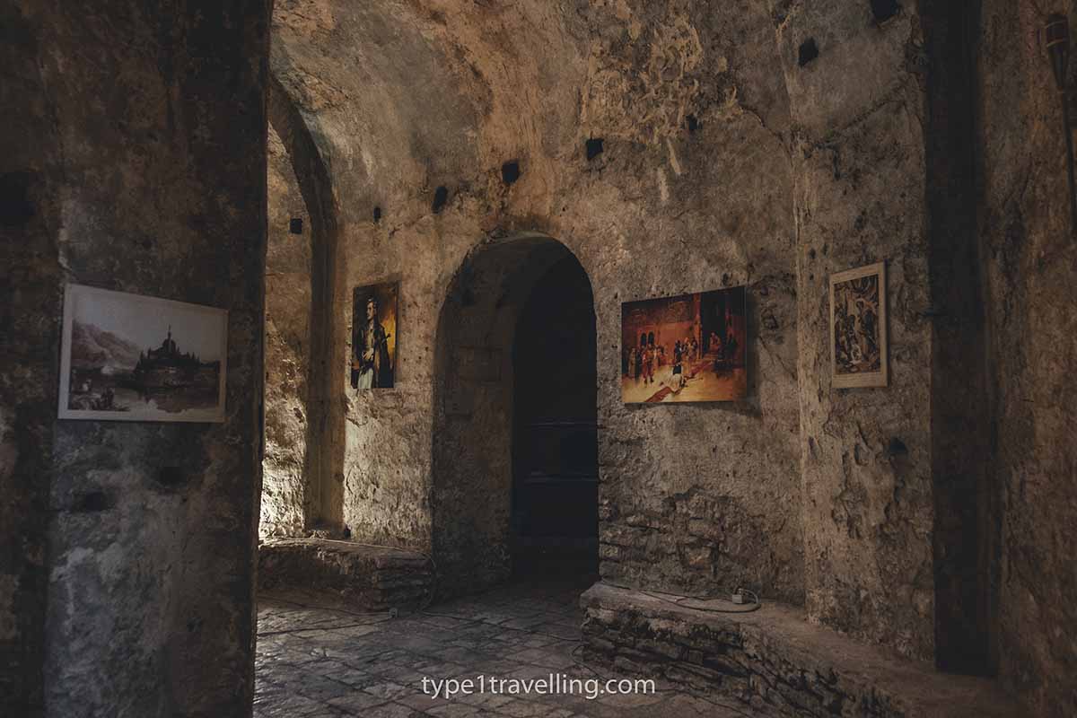 Four paintings hanging on the stone wall of the dimly lit Porto Palermo Castle interior and a doorway leading to a different room.