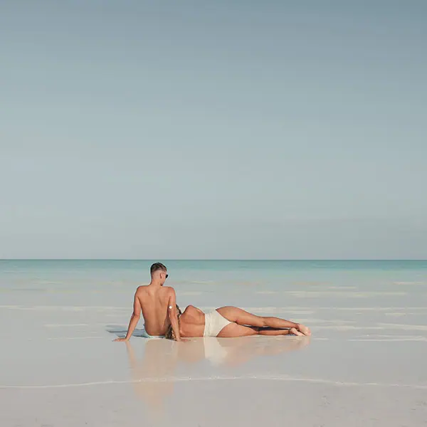 holbox-island A man sitting on the shoreline of a beach while a woman lies next to him with her head resting on his lap.