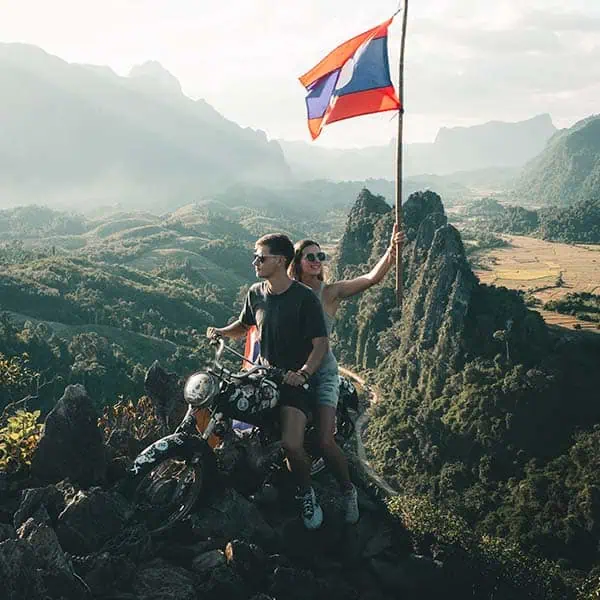 loas-viewpoint A couple sitting on an old motorbike and holding a Laos flag at the top of a view point in Vang Vieng.