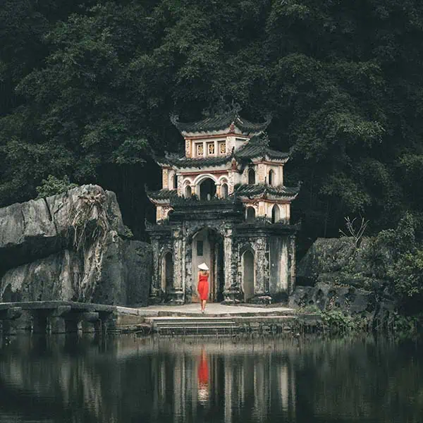tam-coc A woman in a red dress walking towards Bich Dong Pagoda next to a lake.