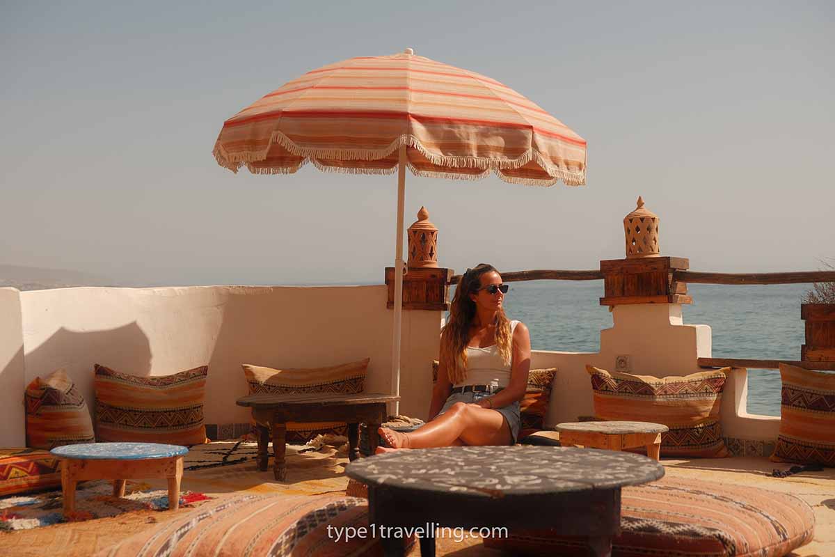 A woman sitting on the floor of a terrace under an orange parasol with the Atlantic Ocean in the background.